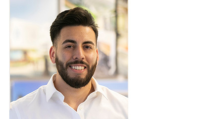 Smiling young man with dark hair and trimmed beard in a white button-up shirt, seated in a bright office with blurred artwork behind him.