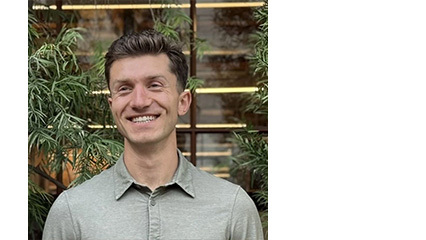 Smiling man with brown hair wearing a light gray collared shirt stands before glass panes, lush indoor plants, and warm lighting.