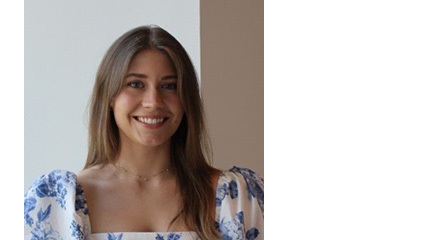 Smiling person with long brown hair, center part wearing a white square-neck dress with blue floral print and puffed sleeves, gold necklace.