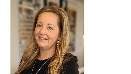 Smiling woman with long wavy blonde hair wearing a black top and dangling earrings, standing in a softly blurred office with shelves.
