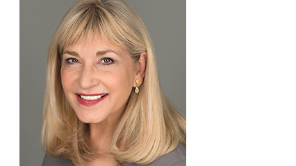 Smiling woman with shoulder-length blonde hair, gold drop earrings and a gray top, headshot on a smooth neutral gray background.