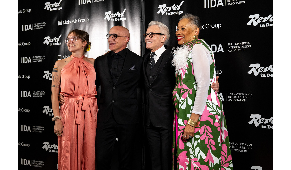 Four smiling people pose on a black backdrop: tattooed woman in coral halter dress, two men in black suits, and woman in green floral dress.