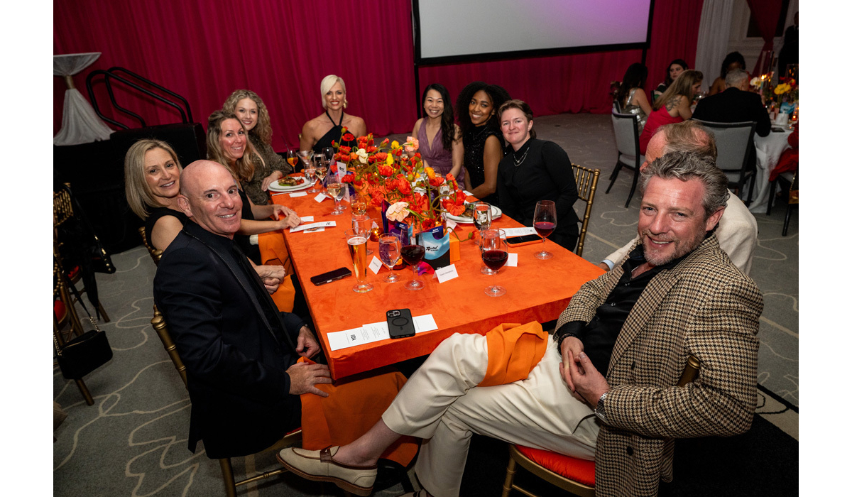 A lively group of well-dressed guests smile around an orange-tablecloth banquet table with bright floral centerpiece and wine glasses.