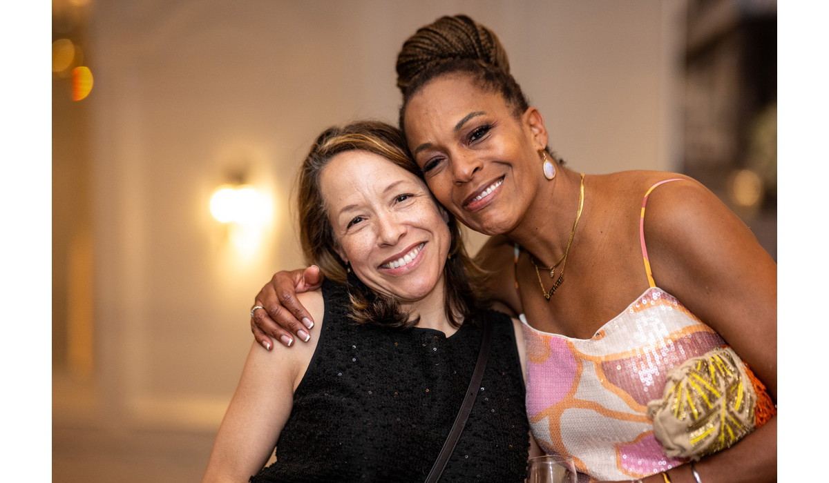 Two women smiling in warm indoor light; one in a black top, the other with braided hair, gold necklace, sequin dress and clutch.