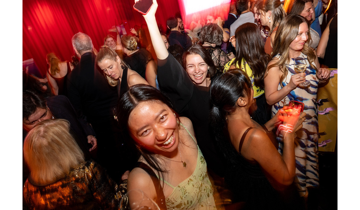 Crowded dance floor, people in dresses laughing and dancing beneath red curtains, one woman raising a phone and another holding wine.