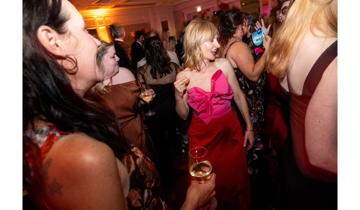 Blonde woman in a pink strapless dress with a large bow dances smiling among crowded guests holding wine glasses in a warmly lit ballroom.