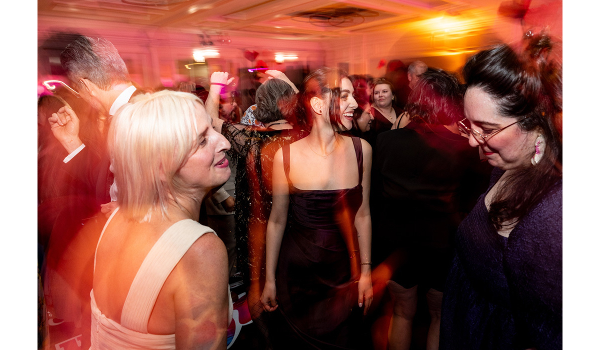 Crowded dance floor under warm amber lights, a joyful laughing woman in a black strapless dress at center, a blonde in white nearby.