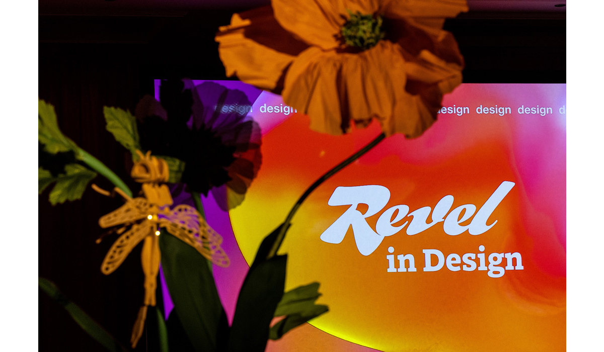 Bold white 'Revel in Design' text overlays a warm orange-pink gradient stage backdrop with oversized paper flowers and a dragonfly ornament.