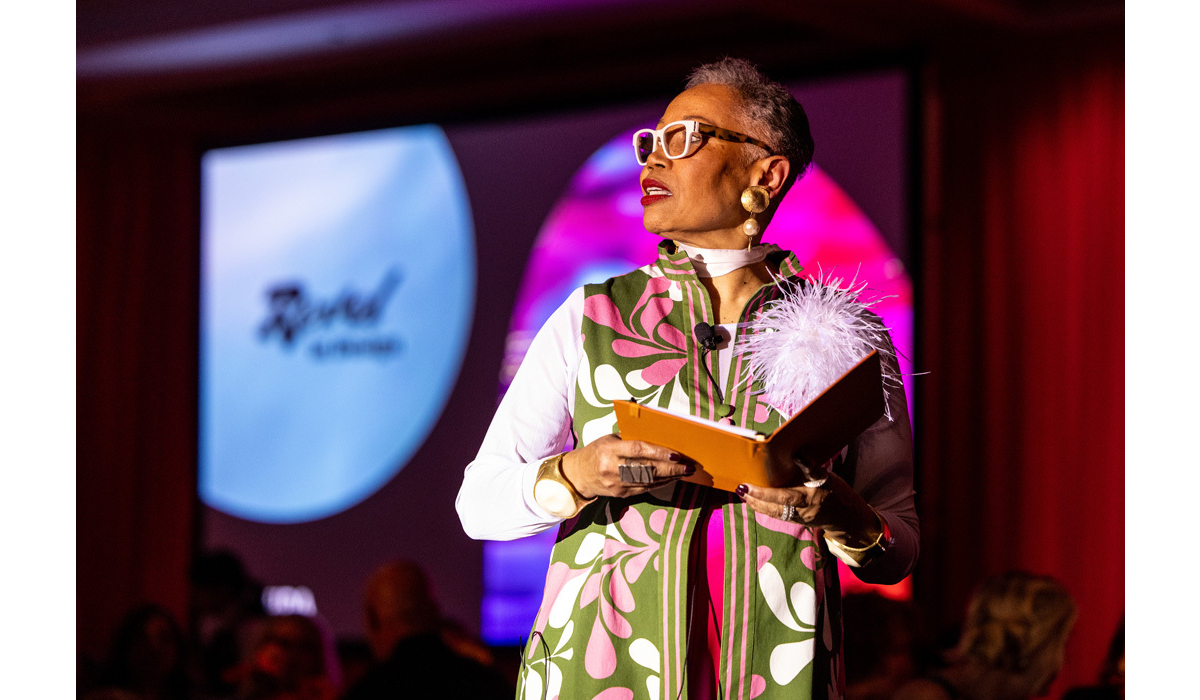 Older woman with white glasses and gold earrings on stage in a green-and-pink vest, holding an orange notebook topped with white feathers.