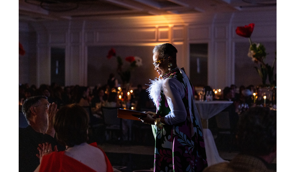 An elegant woman in a colorful floral dress with a white feather corsage reads from a folder while smiling at a candlelit banquet audience.