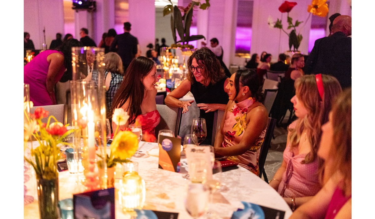 Three women in colorful dresses laugh and chat across a candlelit banquet table adorned with flowers, wine glasses, menus, and pink lights.