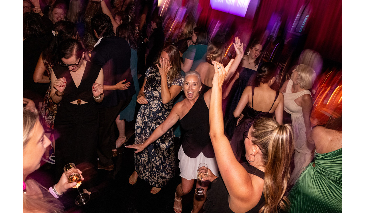 Crowded dance floor of women in dresses dancing lively; a smiling woman in a one-shoulder black top and white skirt raises an arm.
