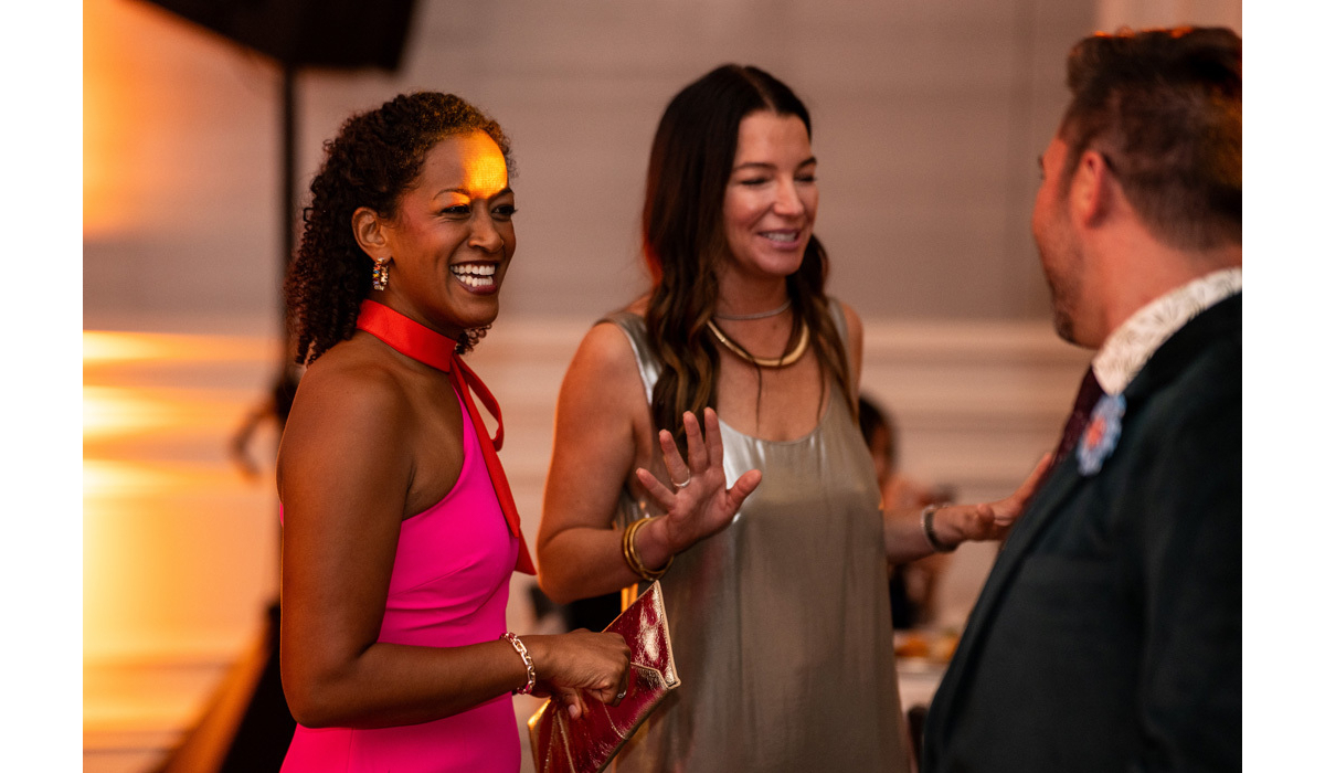 Two women at a reception—one in a hot-pink halter holding a metallic clutch, the other in a silver slip, laughing with an animated man.