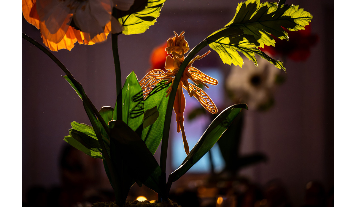 Backlit orange fairy ornament perched among translucent green leaves and orange petals, casting lacy shadows against dim indoor background.
