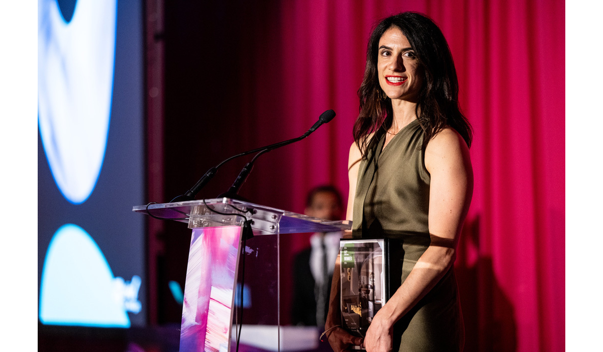 Smiling woman in an olive dress stands at a clear podium holding a glass award, speaking into a microphone against magenta curtains.