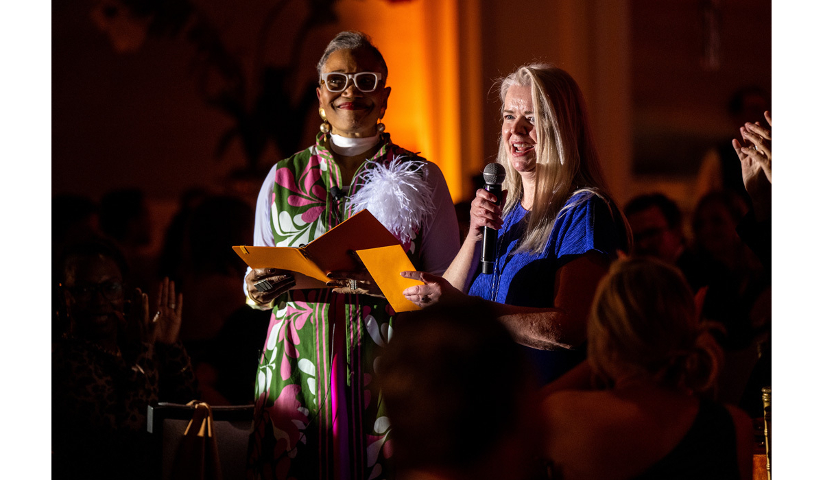 Two women in warm spotlight: a Black woman in white glasses and floral dress holds a yellow folder while a blonde woman speaks into a mic.