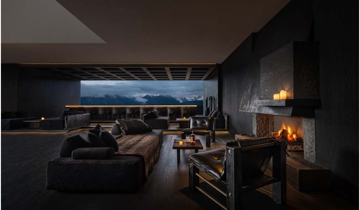 A cozy modern lounge features dark furnishings, a fireplace, and expansive windows showcasing a mountain view under a cloudy sky.