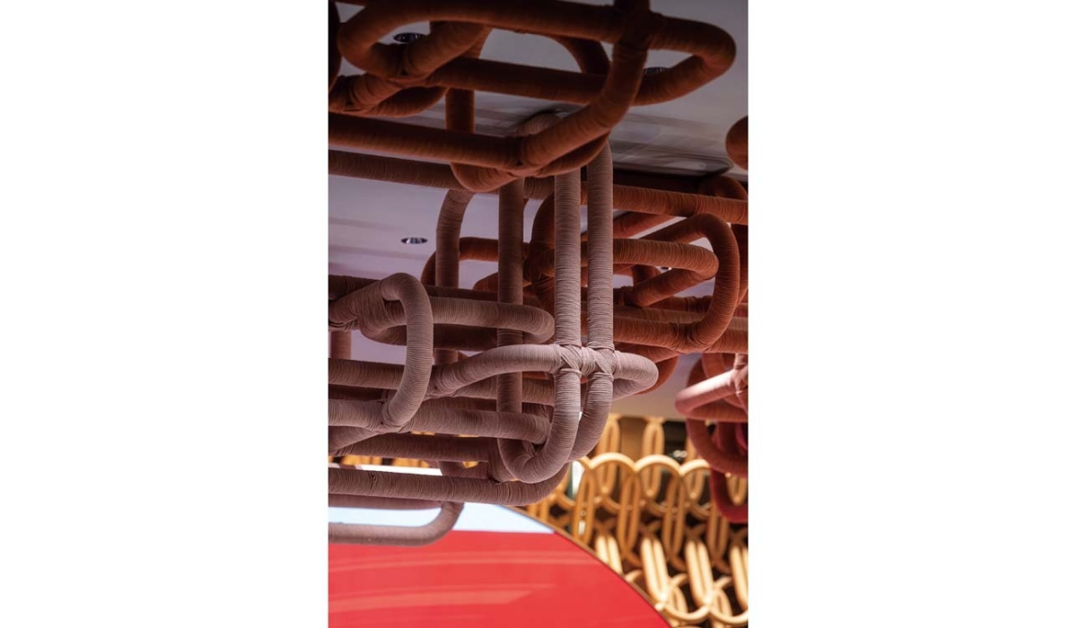 Suspended sculptural ceiling of thick fabric-wrapped tubular loops in terracotta and beige, knotted and weaving above a red curved surface.