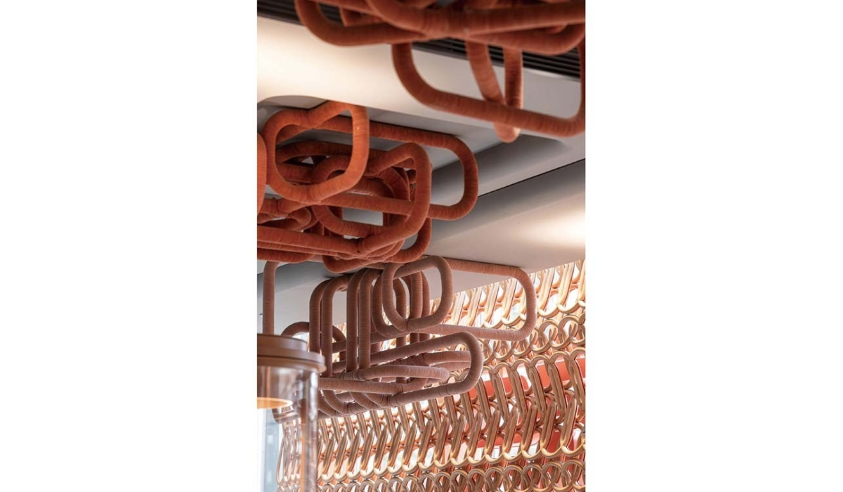 Ceiling installation of interlaced rust-orange fabric-wrapped tubular loops hanging above a repeating geometric copper-toned wall panel.