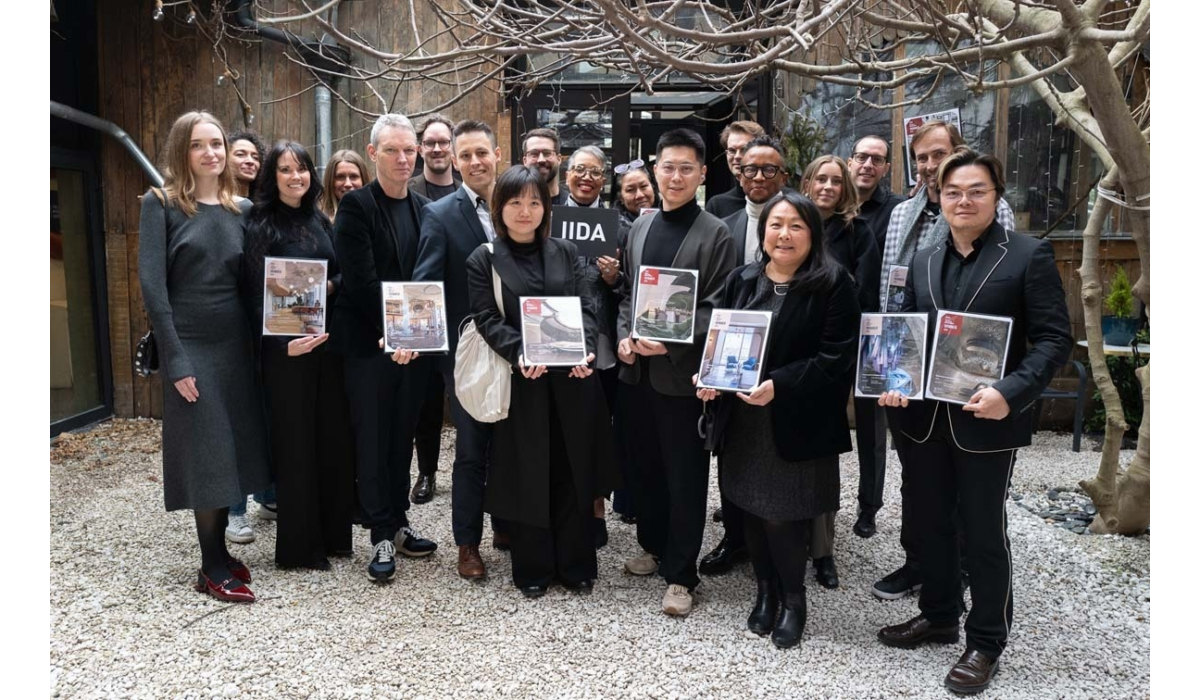 A diverse group of professionals poses outdoors, holding design magazines, in a setting with wooden structures and trees.