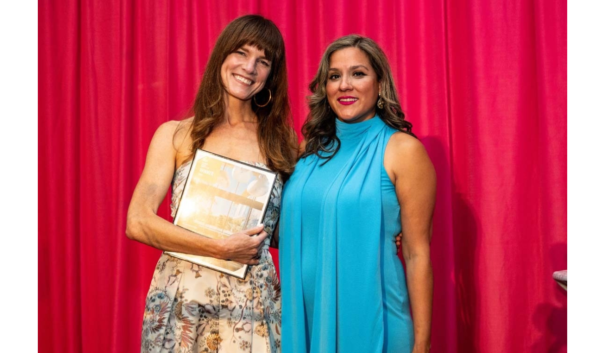 Two smiling women stand before a red curtain; one in a floral dress cradles an award plaque, the other wears a sleeveless turquoise gown.