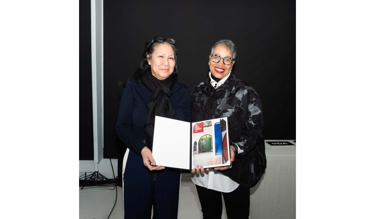 Two women stand together, holding a presentation book showcasing architectural designs, smiling against a dark background.