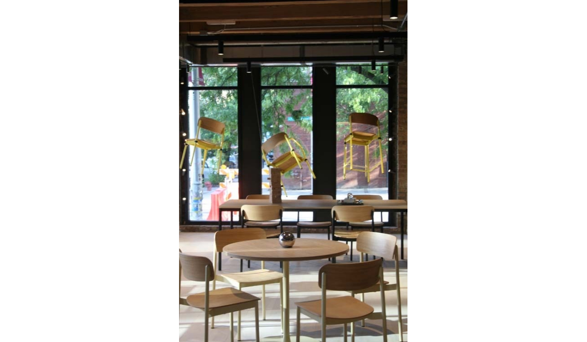 Sunlit café interior with round wooden tables and chairs in foreground and three yellow barstools suspended near floor-to-ceiling windows.