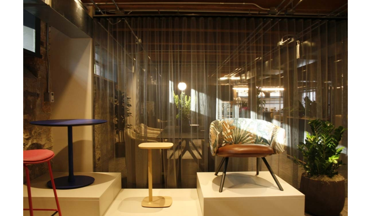 Showroom display with a tropical-print armchair with leather seat on a white pedestal, yellow and blue tables, red stool and potted plants.