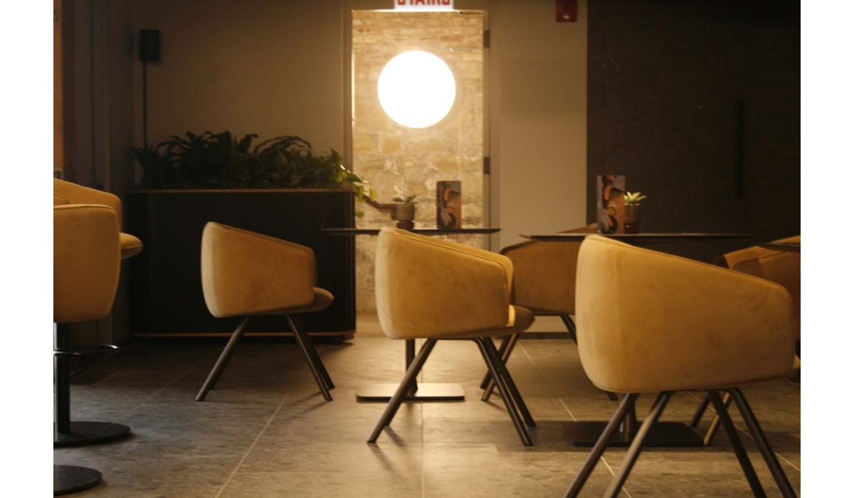 Cozy modern cafe with mustard-yellow upholstered swivel chairs around small round tables, warm circular backlit wall light and low plants.