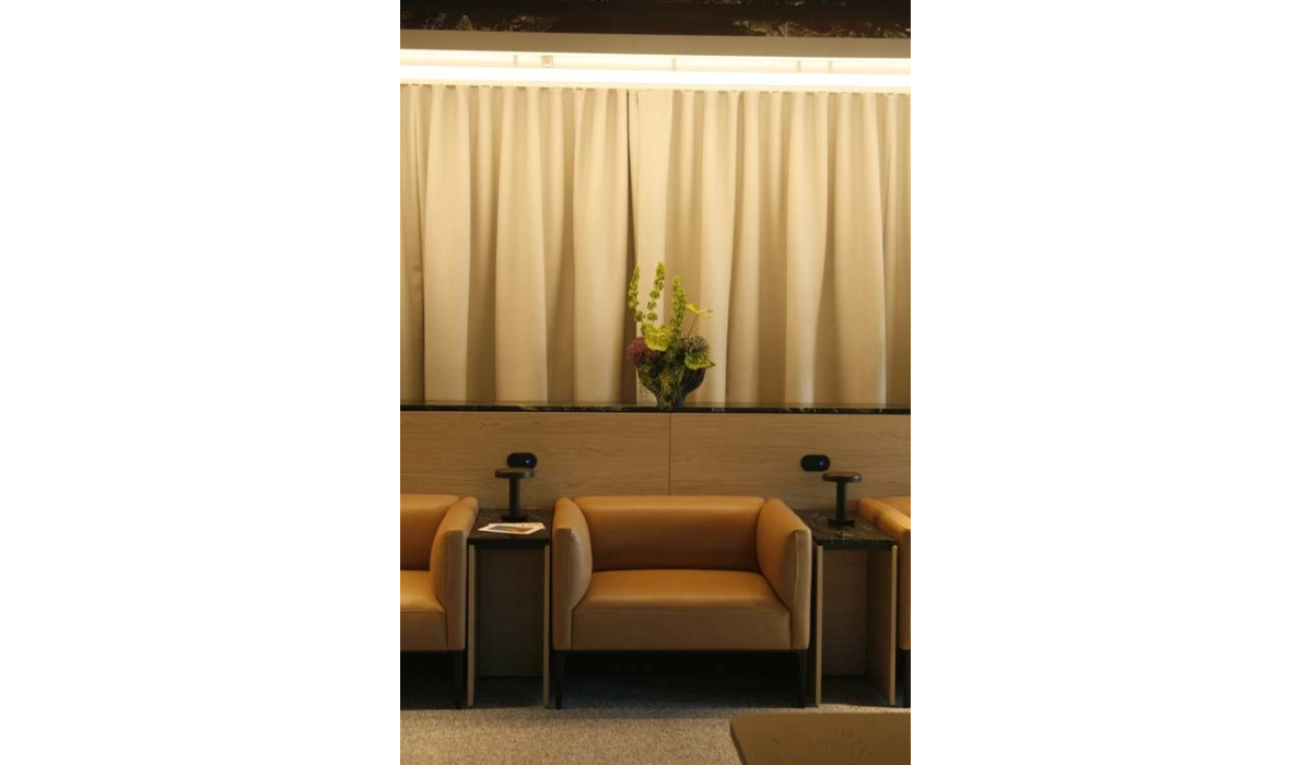 Three caramel leather lounge chairs separated by black marble side tables with lamps sit before beige drapes and a tall floral arrangement.