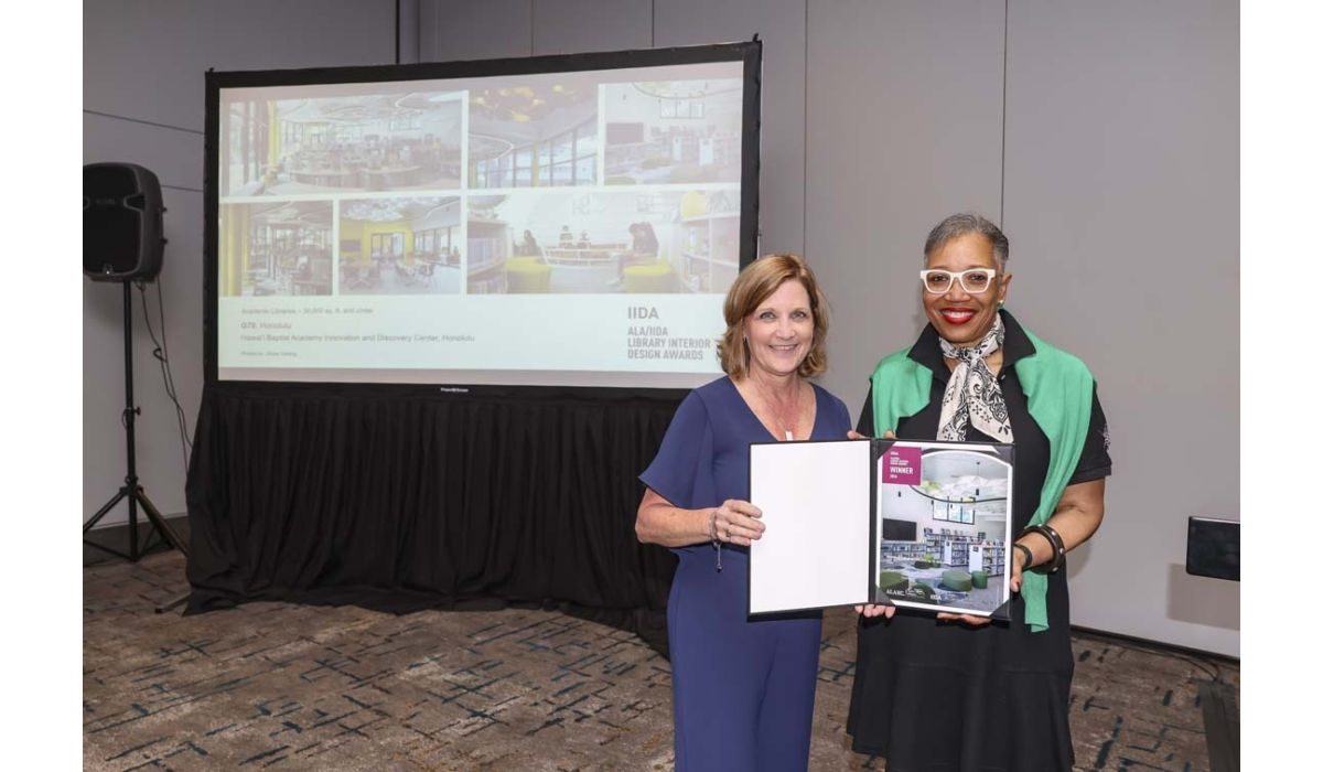 Two smiling women stand in a conference room holding a framed library interior design award, a projection screen visible behind them.