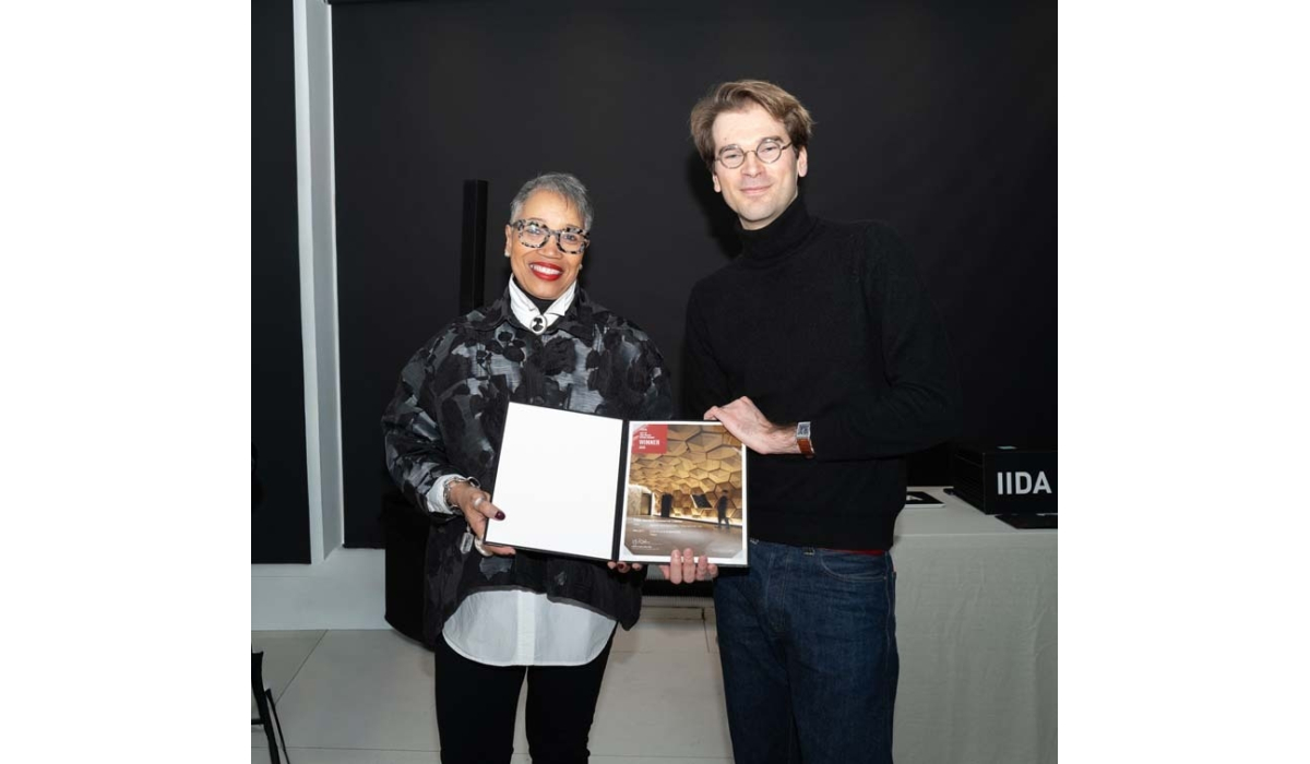 Two individuals stand together, one holding an award document showcasing architectural achievements. A black backdrop emphasizes the moment.