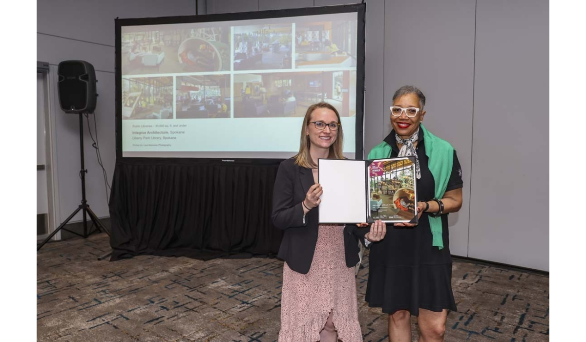 Two smiling women stand in a conference room holding an open award certificate, a projector screen displaying photos behind them.