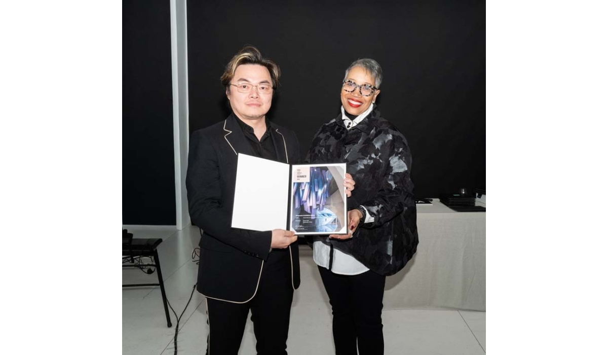 Two individuals stand side by side, each holding an award certificate. One wears a black suit with white accents, while the other dons a stylish black coat.