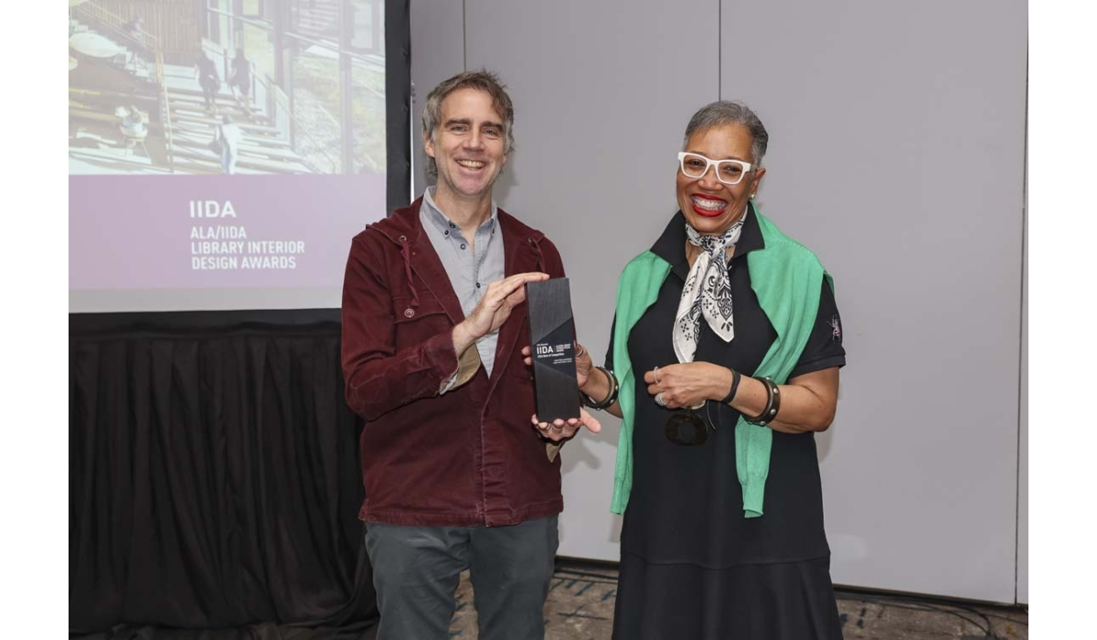 Two smiling people hold a black IIDA Library Design Awards trophy; man in a maroon jacket, woman with white glasses and a green sweater.