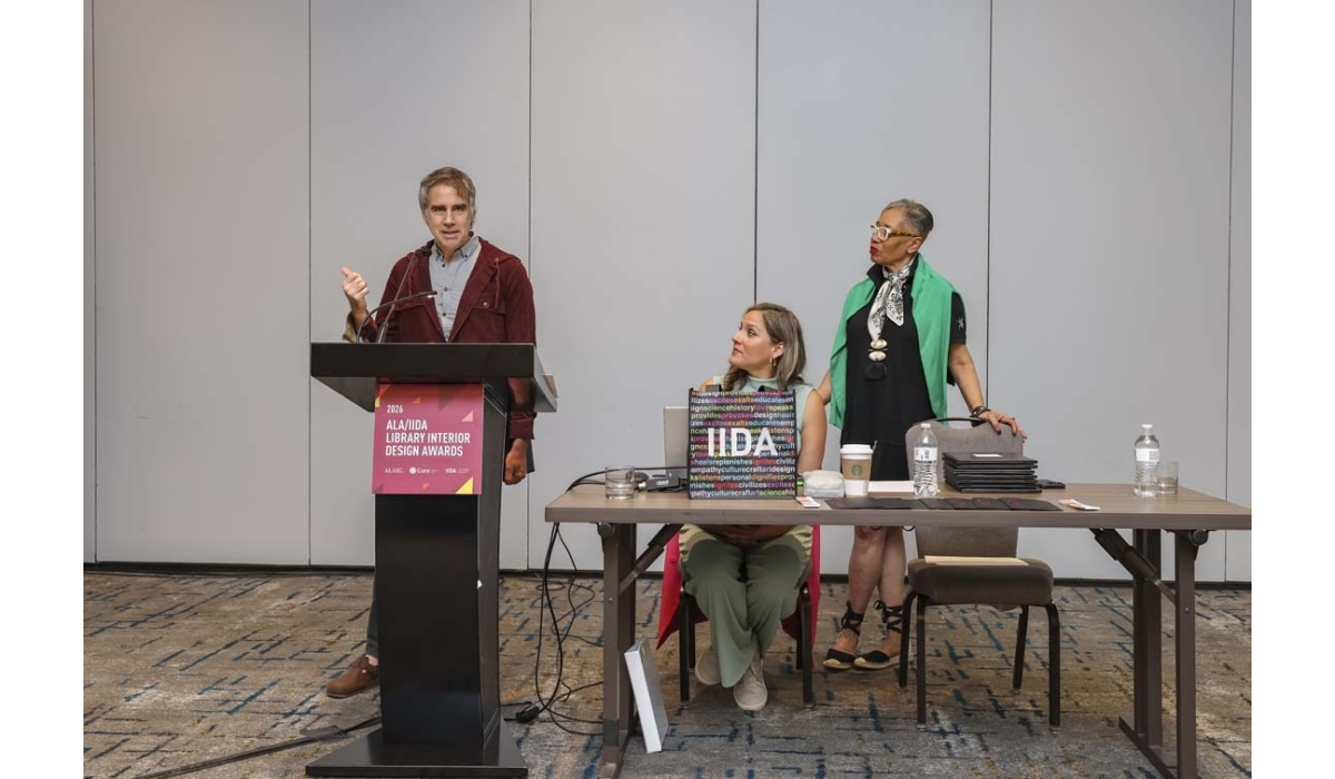 Man at podium speaking at 2026 ALA/IIDA Library Interior Design Awards while two women sit at a table with laptop, coffee and water bottles.