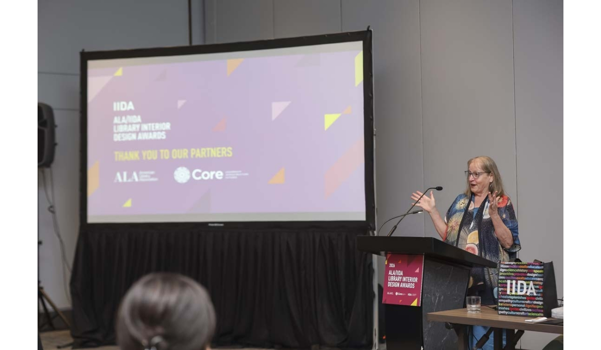 A woman speaker gestures at a podium beside a large purple presentation screen displaying 'Thank you to our partners' and logos.