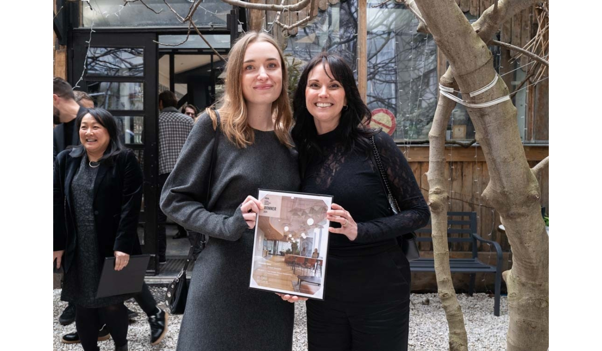Two women stand together, smiling and holding an award certificate, surrounded by a cozy indoor setting with plants and guests.