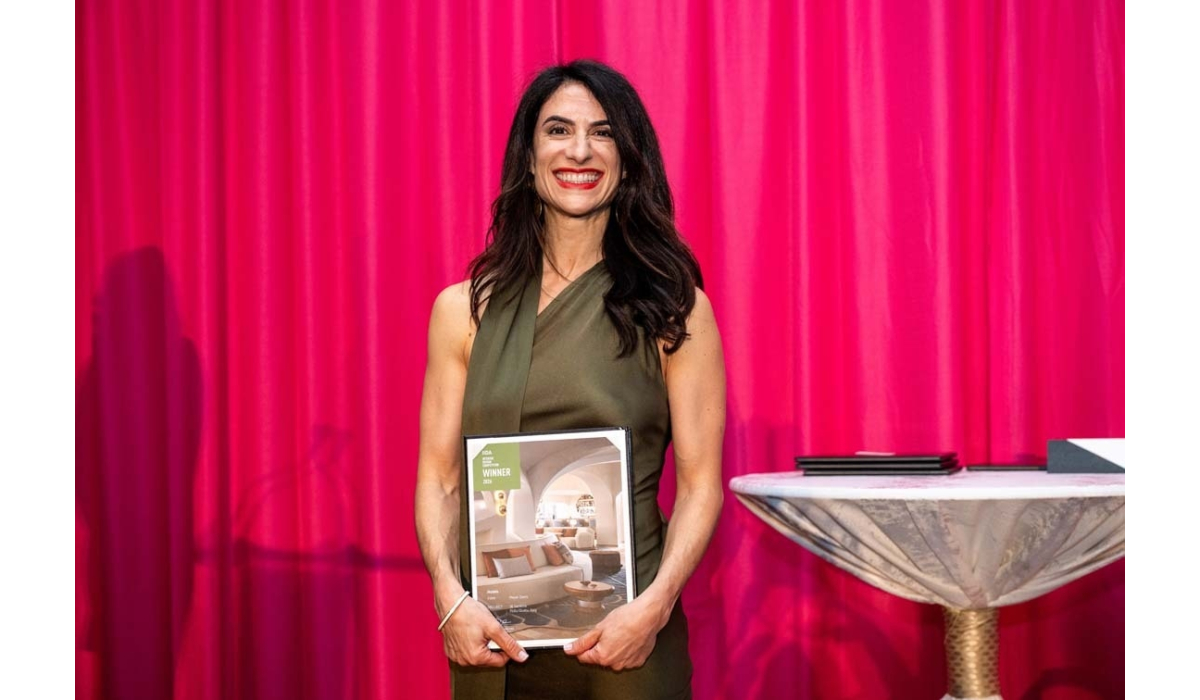 Smiling brunette woman in an olive sleeveless dress holds a framed award booklet labeled WINNER, standing before a bright pink curtain.