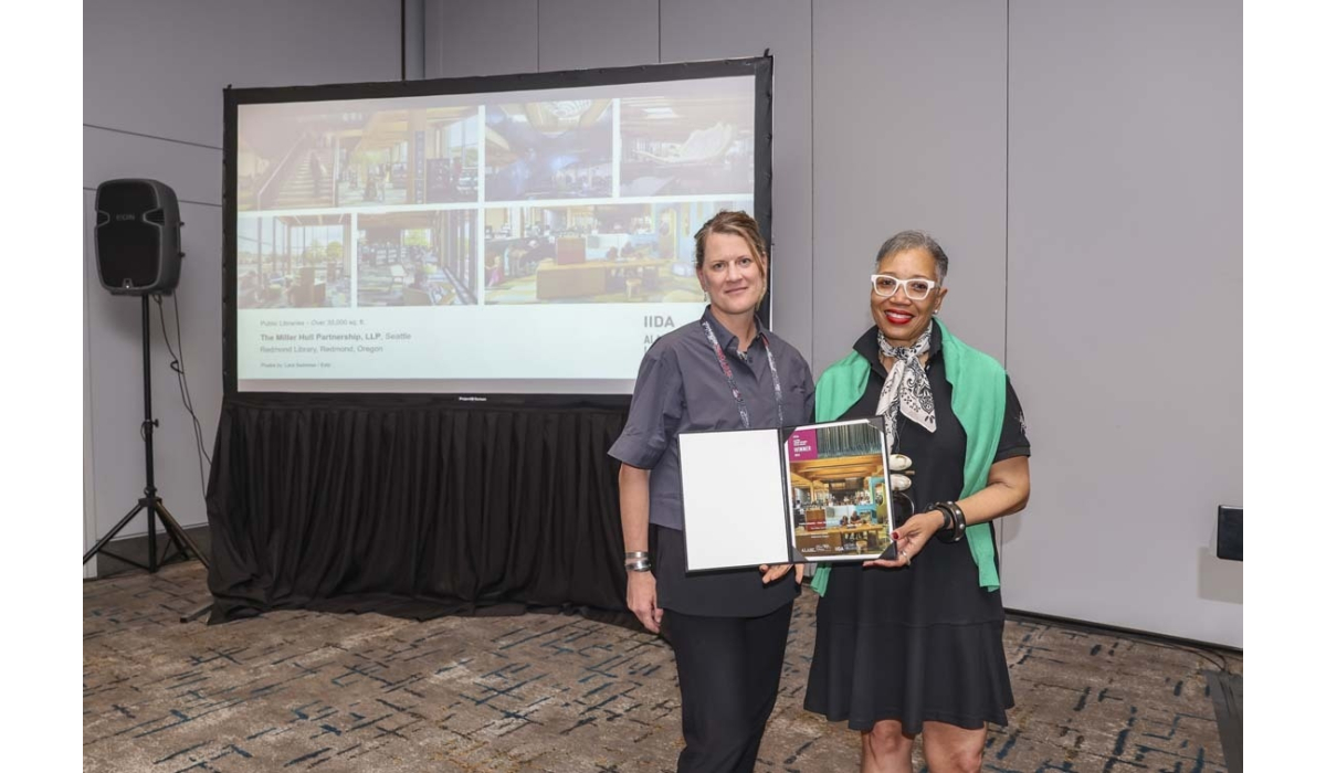 Two smiling women stand in front of a projection screen at a conference, holding an open award portfolio showing colorful library photos.