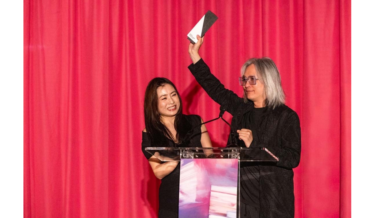Two presenters stand at a clear podium with a pink curtain backdrop; a woman smiles while a man with gray hair and glasses raises an award.