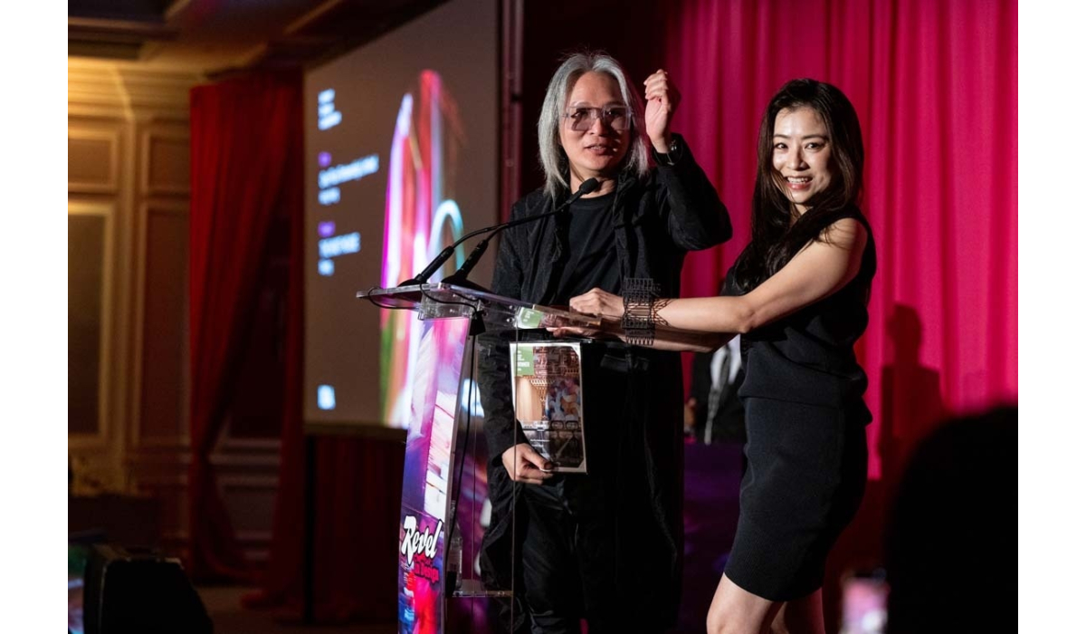A man with gray hair and sunglasses and a smiling woman in a black dress stand at a podium, holding an award against pink curtains.