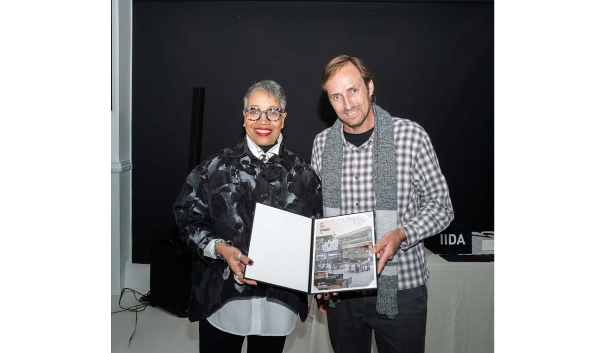 Two individuals stand together, holding an awards booklet, smiling at the camera. A dark background and tables are visible behind them.