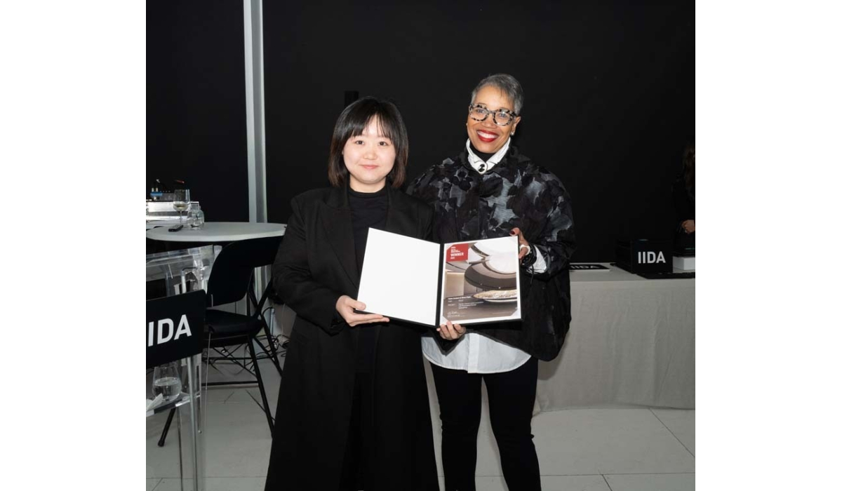 Two women stand together, smiling and holding an award booklet, against a dark backdrop, with a modern table and chairs in the background.