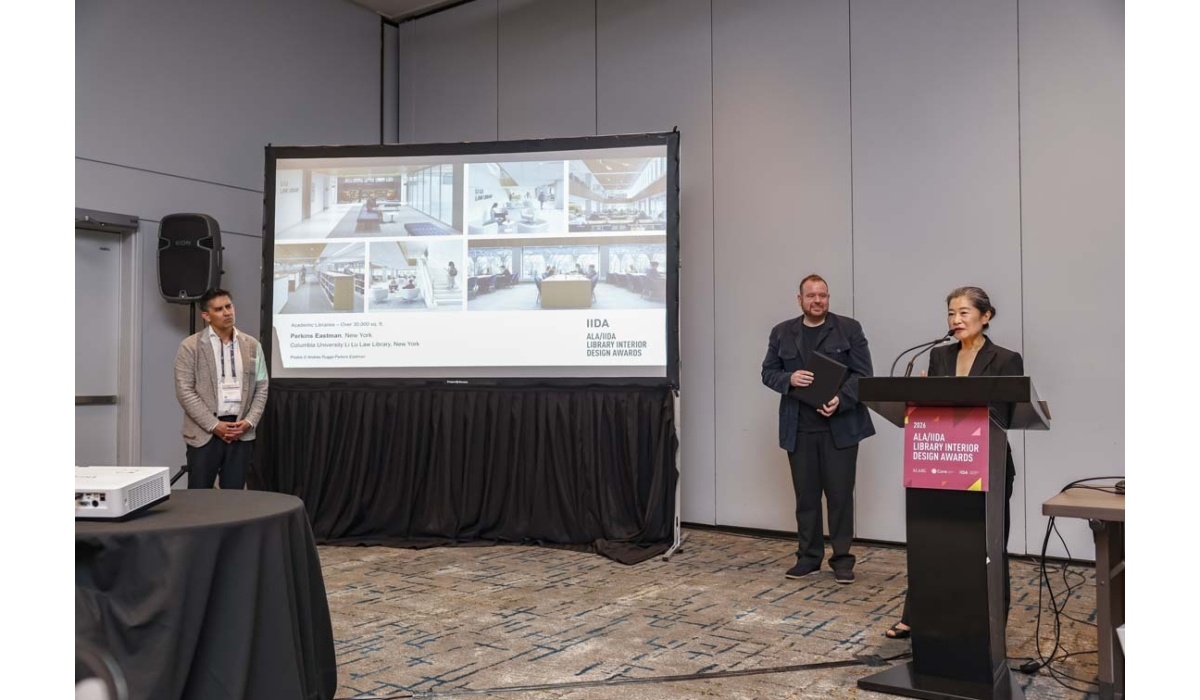 Woman at a podium announces ALA/IIDA library design awards while two men stand near a projection showing library interior photos.