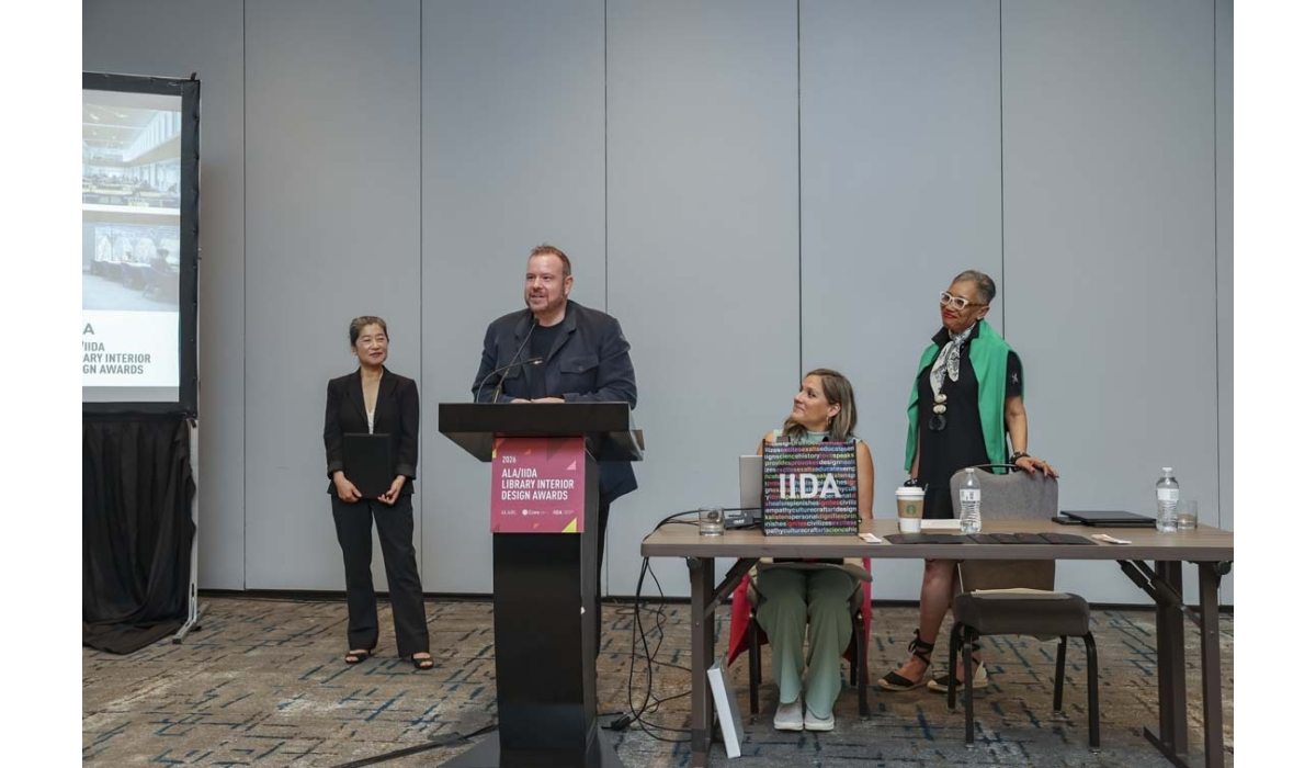 Man speaks at podium for ALA/IIIDA Library Interior Design Awards while three women stand and sit beside him, one holding a colorful plaque.
