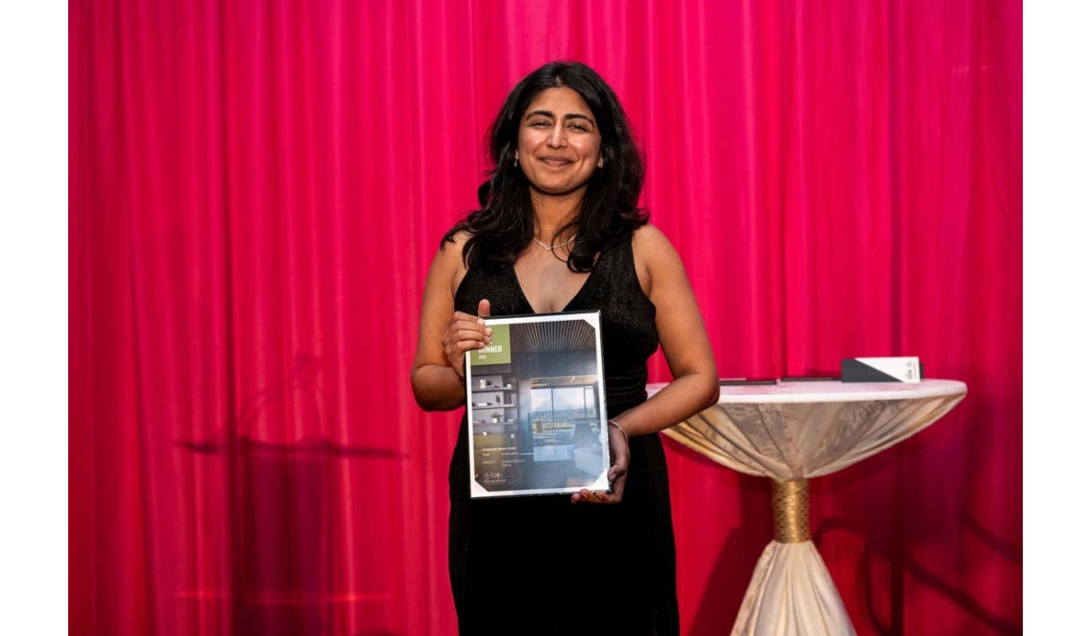 Woman in a black sleeveless dress smiles while holding an award plaque before a bright fuchsia curtain beside a small draped table.