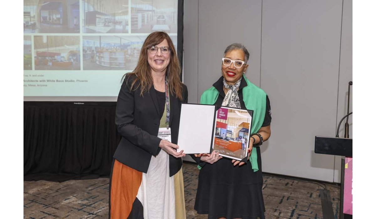 Two smiling women at a conference hold an award plaque proudly; one wears white glasses and a green shawl, the other a black blazer.