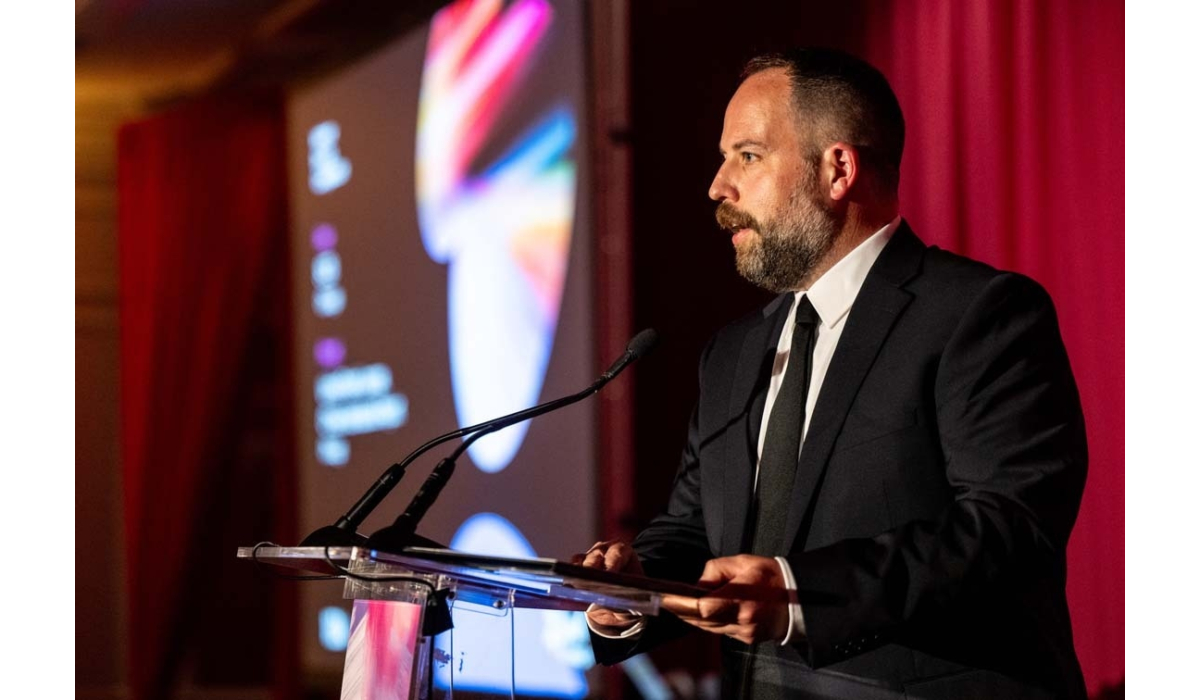 Bearded man in a black suit and tie speaks at a clear lectern with dual microphones, colorful projection and red curtains in background.