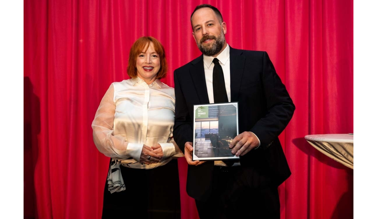 Smiling red-haired woman in a cream satin blouse stands with a bearded man in a black suit holding a framed 'Winner' award, red curtain.
